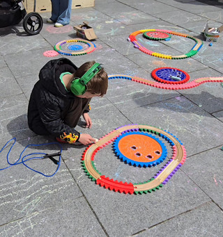 We Invented The Weekend Festival - Boy in green headphones crouches on pavement, arranging colorful circular gear tracks and chalk drawings in a playful outdoor scene