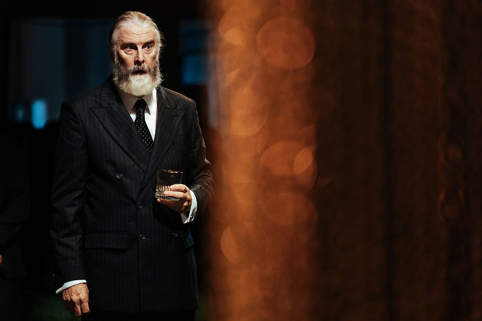 David Threlfall as King Lear wearing a dark pinstripe suit and holding a glass during the Royal Exchange Theatre's contemporary production of Shakespeare's tragedy.