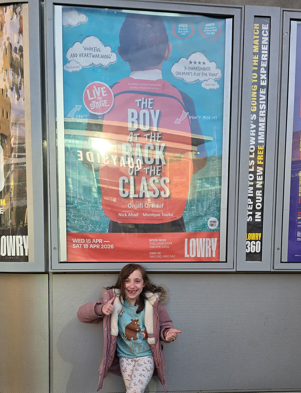 A girl smiles and gestures enthusiastically in front of a theater poster for "The Boy at the Back of the Class" at The Lowry. Bright and colorful.
