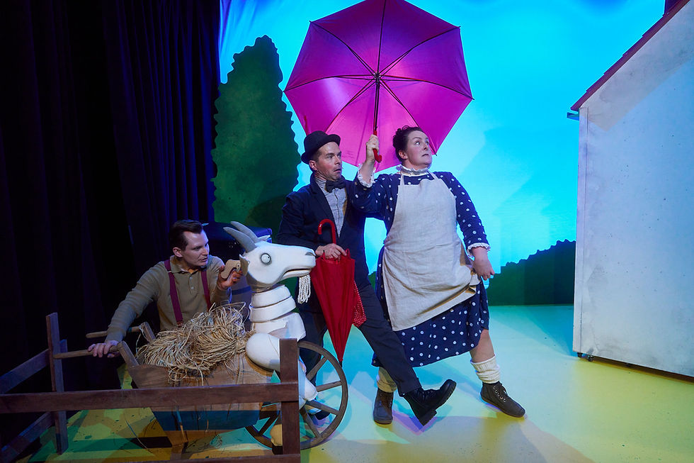 from the stage adaptation of A Squash And a Squeeze, three actors on stage; one with a polka dot dress holds a pink umbrella. Another pushes a goat cart. Colourful tree backdrop.