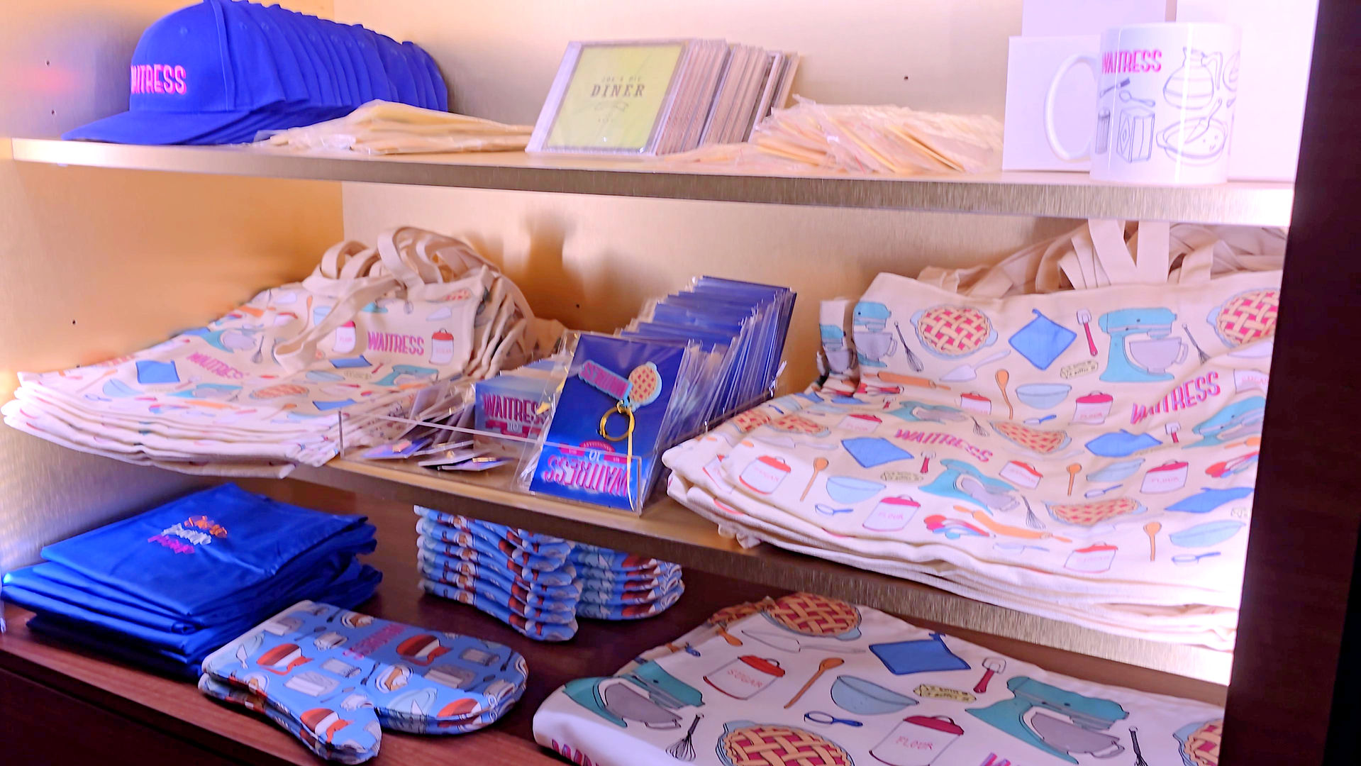 Shelves filled with Waitress The Musical merchandise including tote bags, oven mitts, mugs, keyrings, caps, tea towels and CDs displayed at the theatre merch stand.