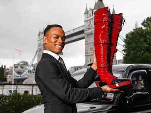 Man holding shiny red boots, smiling near a black taxi, with Tower Bridge in the background on a cloudy day.
