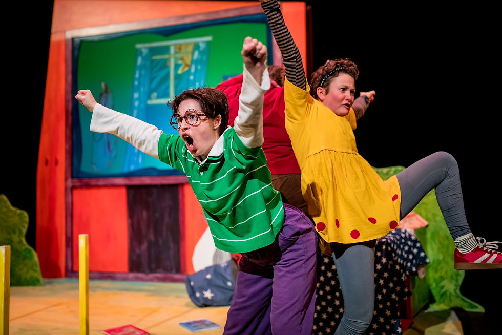 Two actors in colorful costumes energetically pose on stage. One in green and purple, the other in yellow with red spots. Theater set behind.