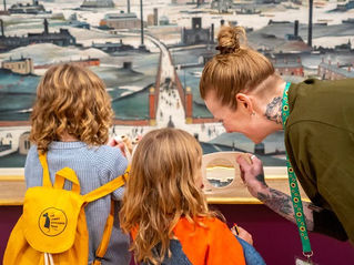 An adult wearing a sunflower lanyard helps two children explore an artwork in the LS Lowry Collection using a magnifying glass at Lowry.