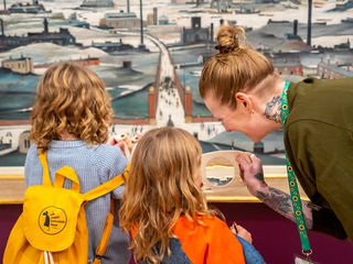 An adult wearing a sunflower lanyard helps two children explore an artwork in the LS Lowry Collection using a magnifying glass at Lowry.