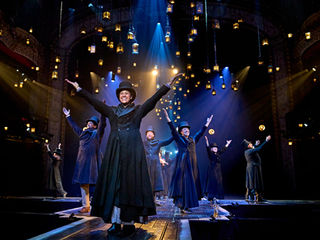 The Old Vic’s production of A Christmas Carol - Performers in dark coats and top hats dance energetically on a stage lit by hanging lanterns and blue spotlights, conveying joy and elegance.
