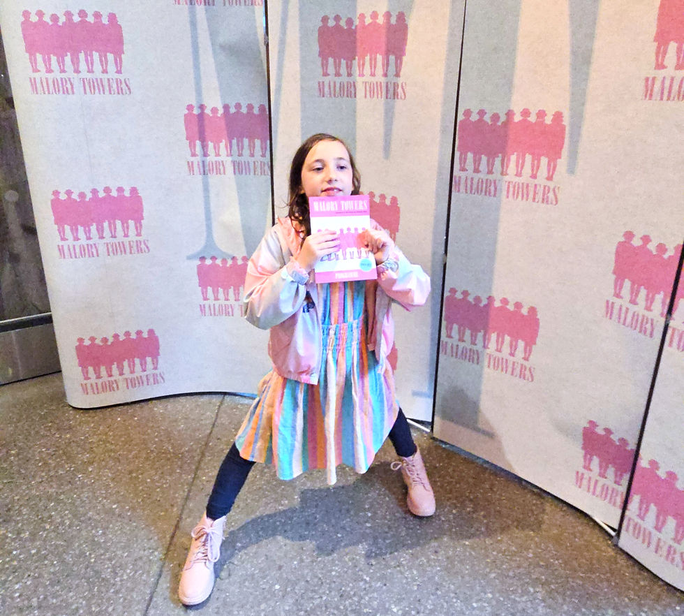 Girl posing with a Malory Towers book in front of a pink Malory Towers backdrop, wearing a pastel striped dress and jacket.