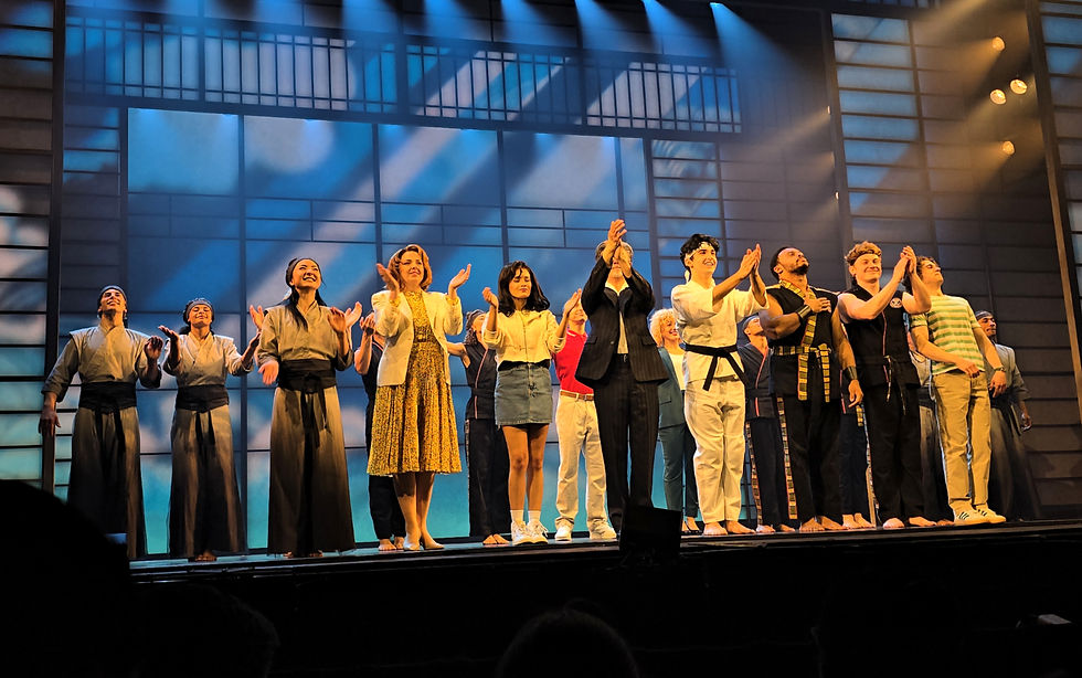 Performers in varied costumes stand in a line on stage, clapping, against a backdrop of blue-lit windows, creating a celebratory mood.