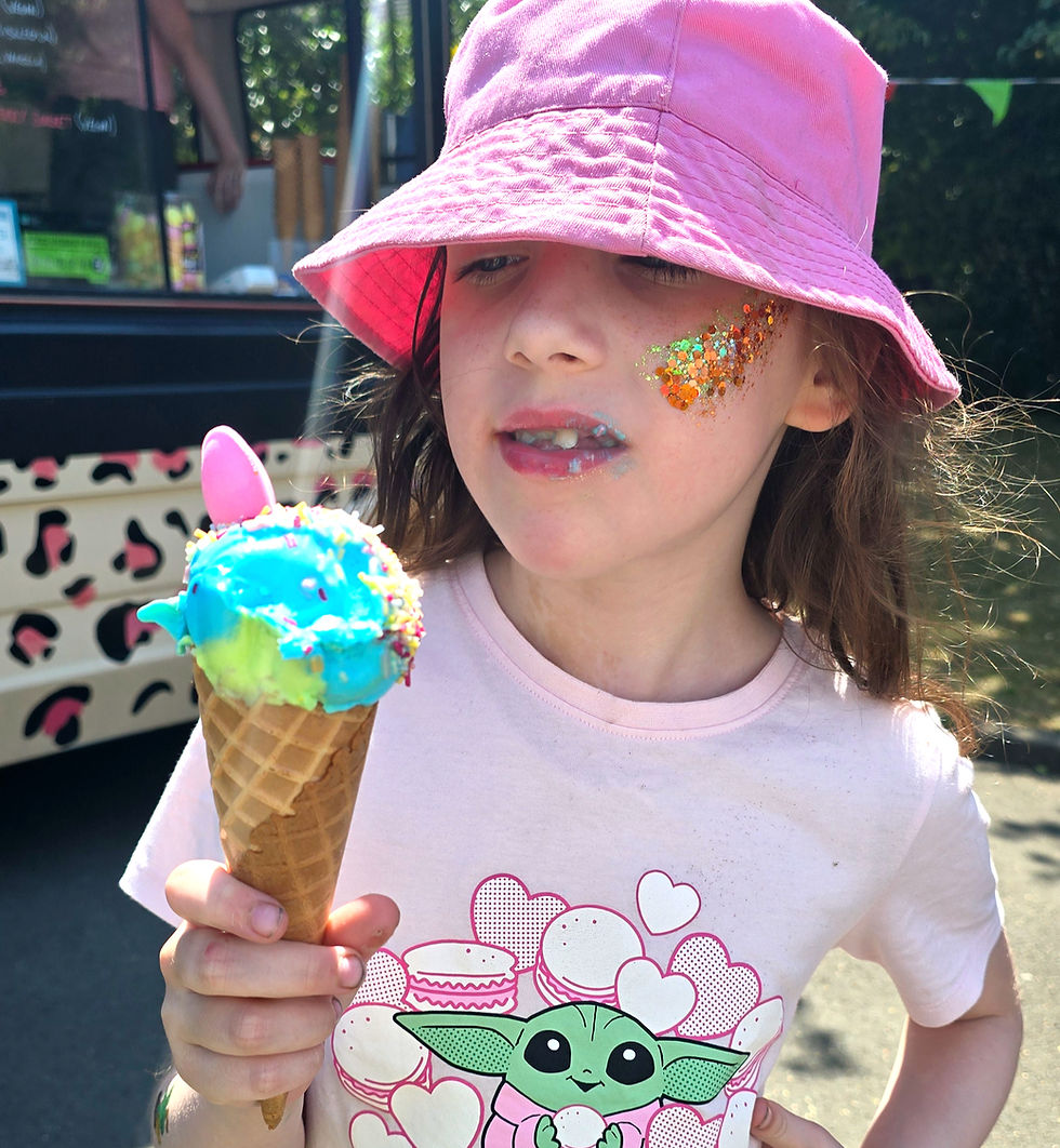 Girl in pink hat and Grogu shirt holds a rainbow ice cream cone, with glitter face paint at an outdoor stand.