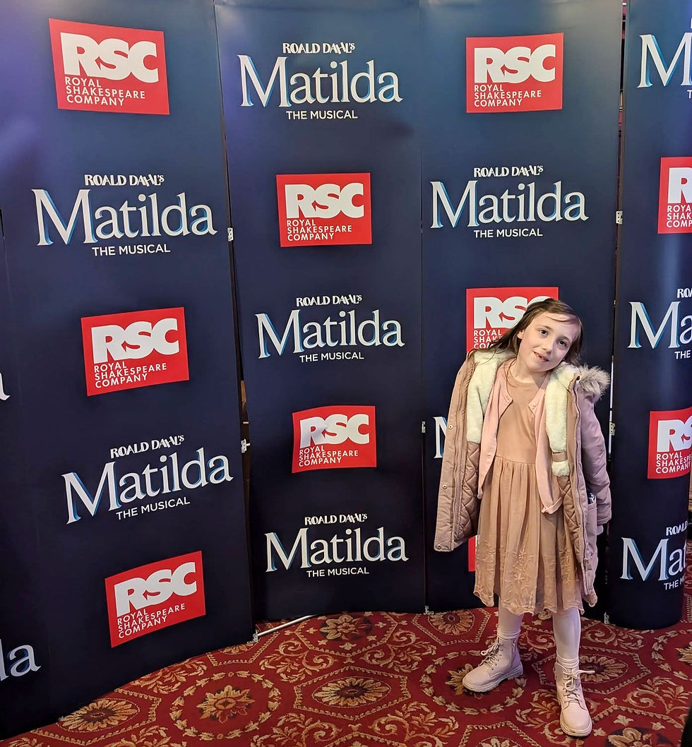 Girl posing in front of "Matilda The Musical" and "RSC" branded banners. She wears a pink dress and coat. Red carpet visible below.