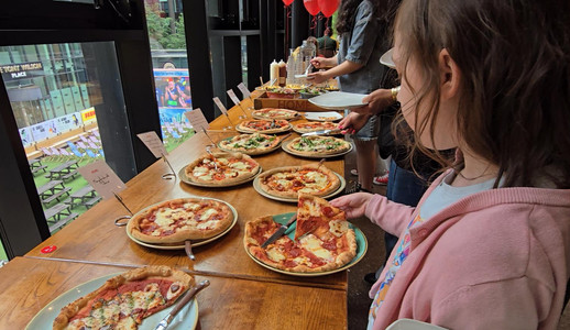 HOME Manchester - Girl in pink hoodie serves pizza at an indoor buffet table, with multiple pizzas, salads, and a city view through large windows.