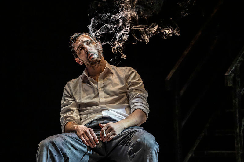 Ralf Little as Alec Leamas sits smoking beneath dramatic lighting during a scene from The Spy Who Came in from the Cold.
