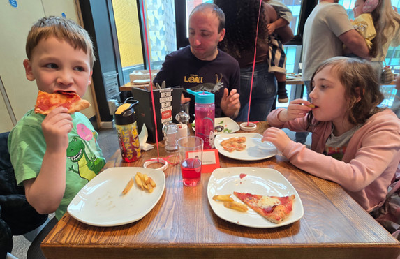 HOME Manchester - Kids and a man eat pizza at a restaurant table, with drinks and plates, while families gather in the background.