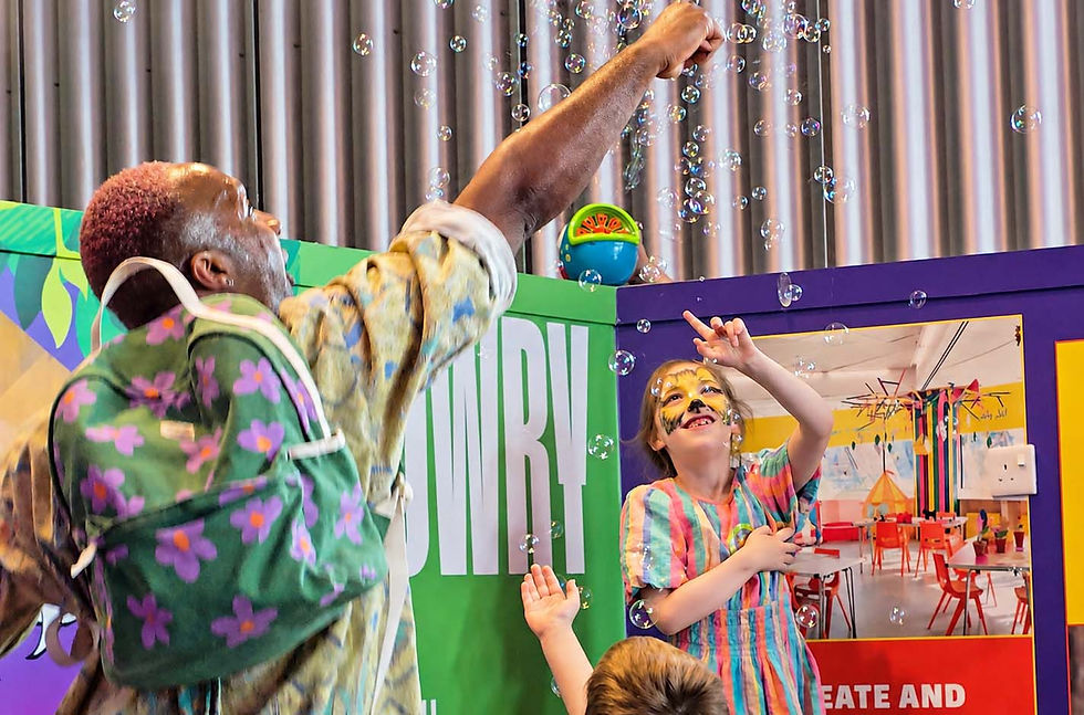 Man and children reach for bubbles in a colorful indoor exhibit; girl with face paint smiles.