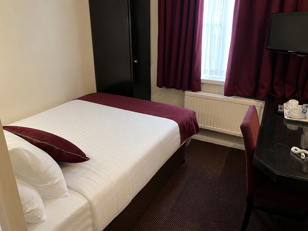 Small hotel room with a made bed, burgundy curtains, desk, TV, and tea tray; calm, tidy interior with daylight from the window