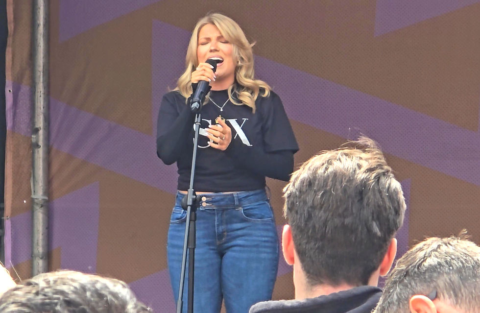 Blonde woman sings into a microphone on an outdoor stage, wearing a black shirt and jeans, with audience heads in foreground.