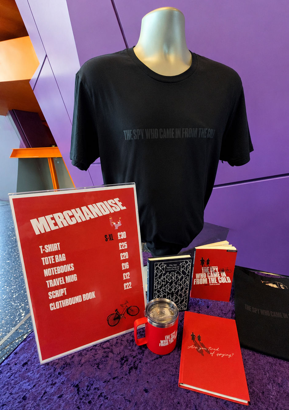 Merch display with black Spy Who Came in from the Cold T-shirt, red price sign, mug, books, and tote on purple backdrop.