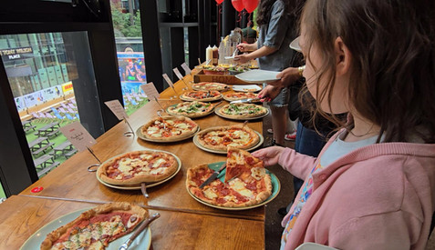 HOME Manchester -  Girl in pink hoodie serves pizza at an indoor buffet table, with multiple pizzas, salads, and a city view through large windows.