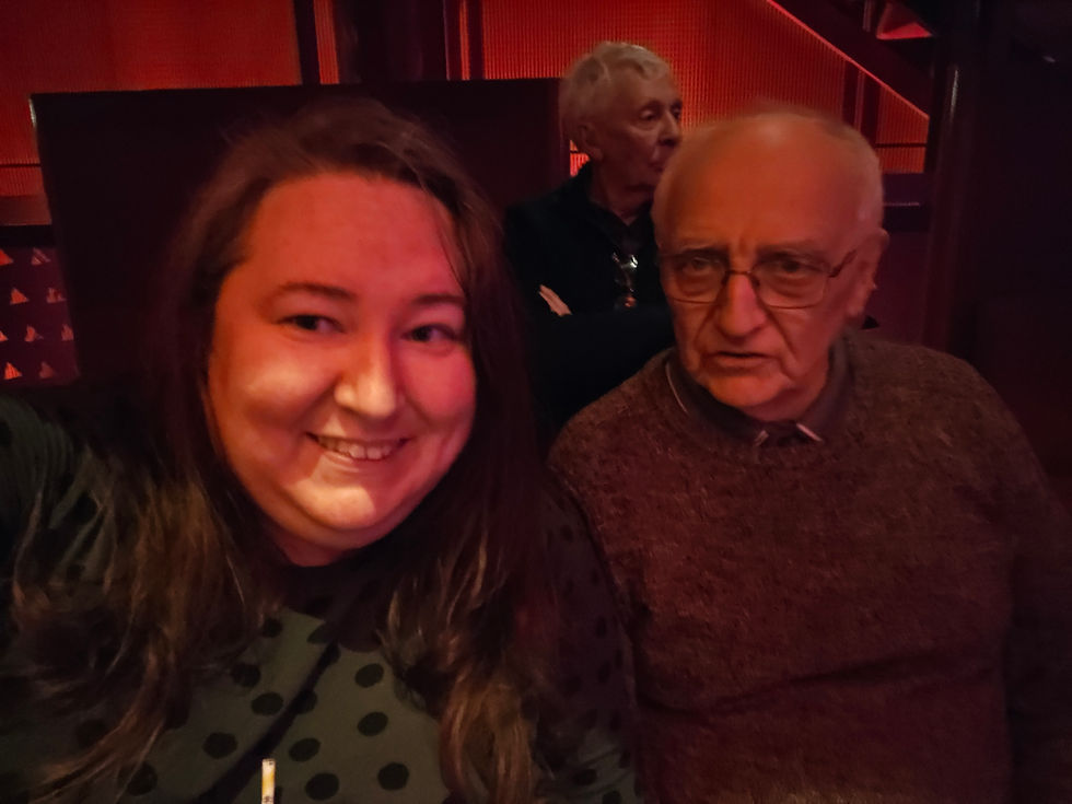 Two people sit in a dimly lit setting with red hues. One smiles warmly, the other appears thoughtful. A third person is partially visible.