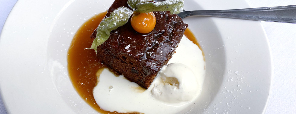 Sticky toffee pudding topped with a physalis garnish, served with cream and toffee sauce in a white bowl.