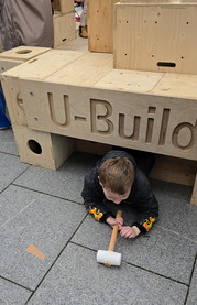 We Invented The Weekend Festival - Boy crawls under a wooden U-Build structure, holding a mallet on gray pavement in a playful, focused scene.