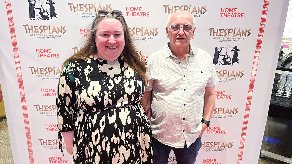 Smiling woman and older man pose before a Thespians Greece the Musical Home Theatre step-and-repeat backdrop.