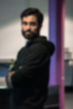 Eben Figueiredo in rehearsals for A Doll’s House. Photo by Chris Saunders. A person with dark hair and a beard stands with their arms folded and looks serious. They wear a black hoodie and look slightly over their left shoulder.