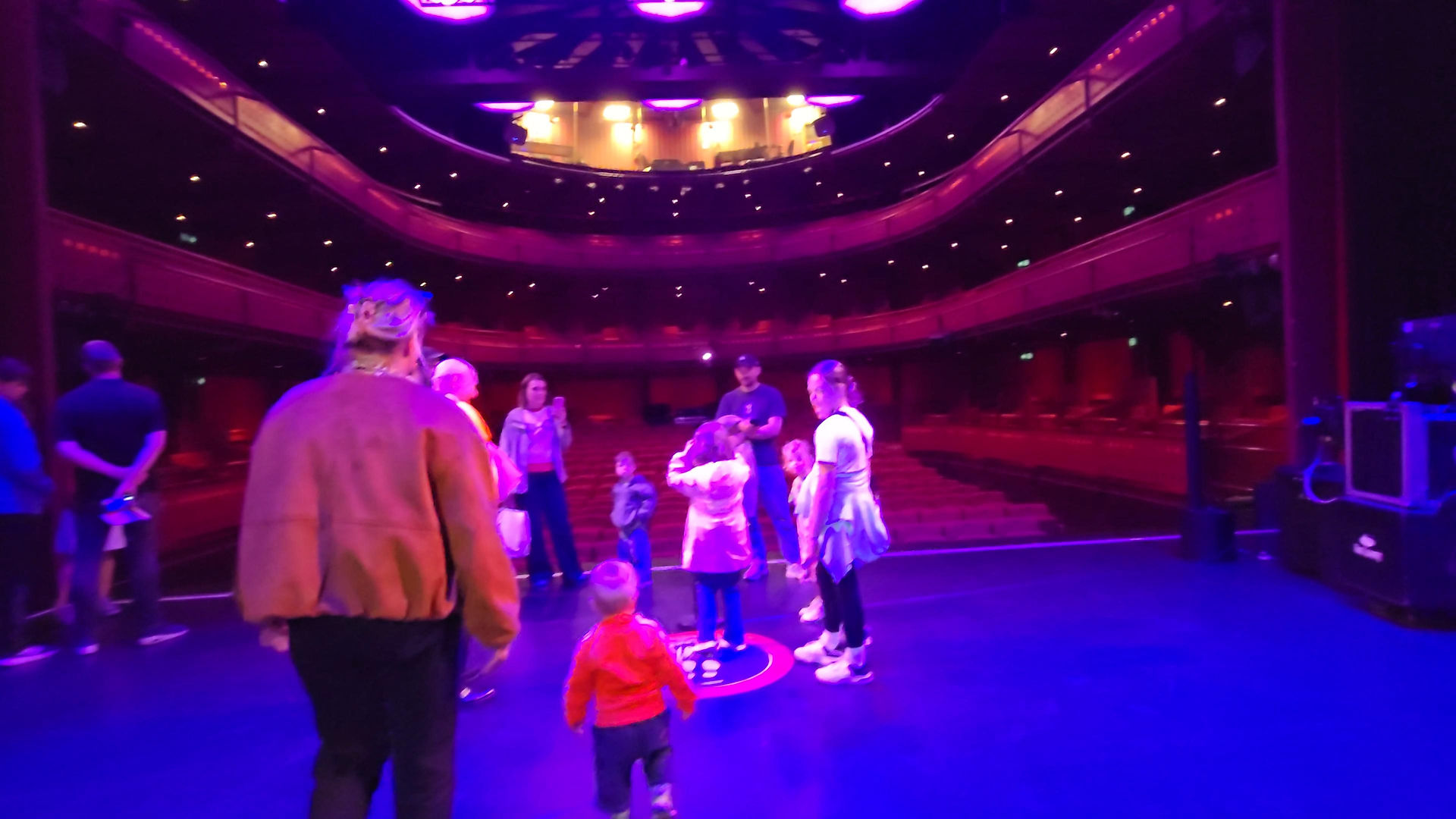 Lowry Backstage Tour -  Group of people and children on a theater stage in a purple-lit auditorium, looking around during a tour.