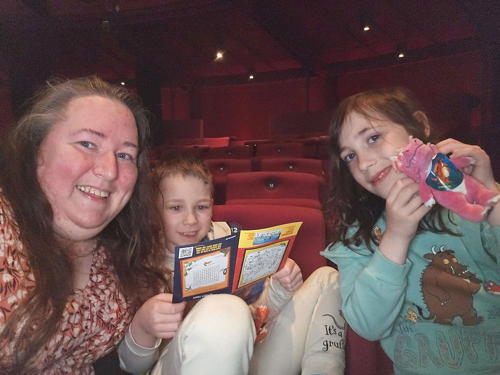 Three people smile in a theater. A child holds a pink stuffed toy and another reads a colorful booklet. Red seats fill the background.
