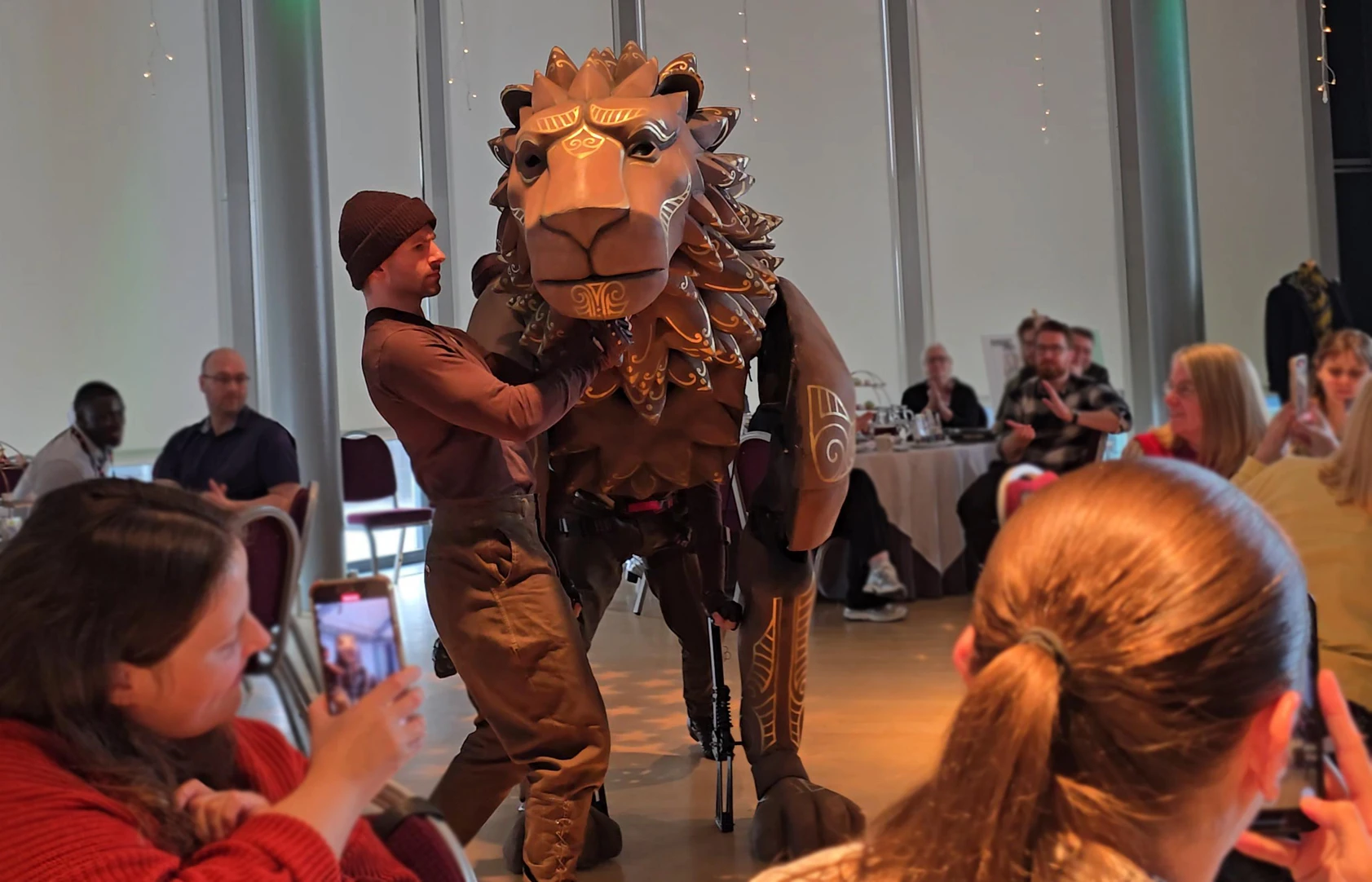 Man in brown costume poses with a large lion mascot at an indoor event while people watch and take photos.