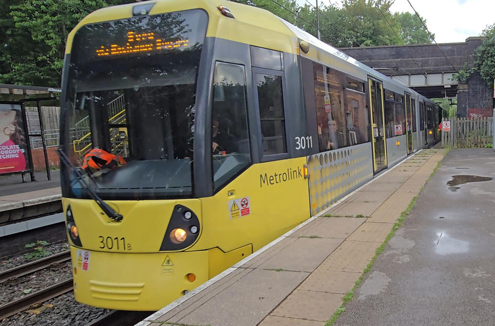 Yellow Metrolink tram 3011 at a leafy station platform, headlights on, with a bridge and wet pavement in the background.