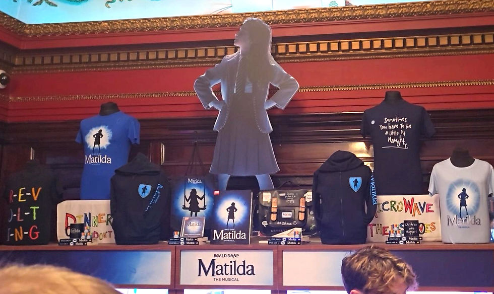 Merchandise display for "Matilda the Musical" with T-shirts, bags, and a cardboard cutout of Matilda. Red and gold ornate background.