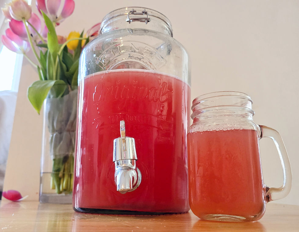 Glass jar with red juice and tap beside matching glass mug; pink tulips in vase in background on wooden surface.