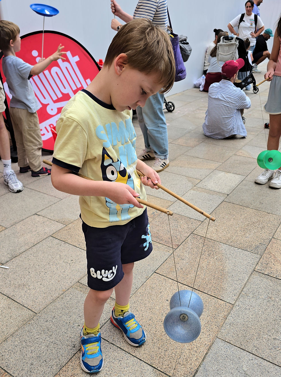 Boy in Bluey shirt plays with a blue diabolo indoors while others watch nearby, with a red sign in the background.