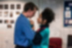 Tom Glenister and Siena Kelly in rehearsals for A Doll’s House. Photo by Chris Saunders. (3). Two people stand closely together, as if about to kiss. One wears a blue shirt and has short blonde hair, the other wears a multi-coloured fleece and has dark curly hair tied up. They clutch each other’s hands between their chests.