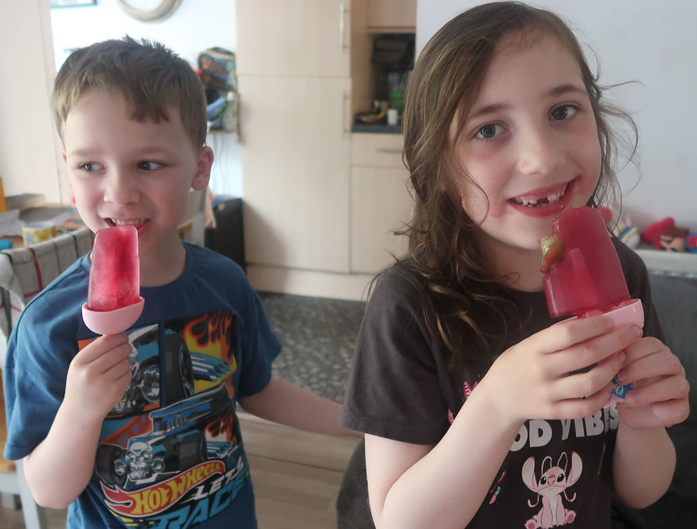 Two children in a kitchen smile while eating red popsicles, the boy in a blue Hot Wheels shirt and the girl in a black Stitch shirt.