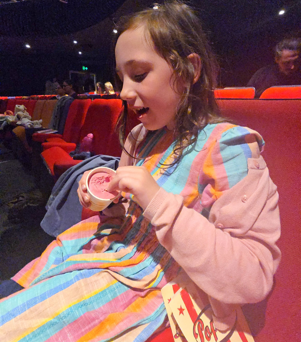 Young girl in a theater seat eats pink ice cream, wearing a colorful striped dress, with red seats and a popcorn box nearby.