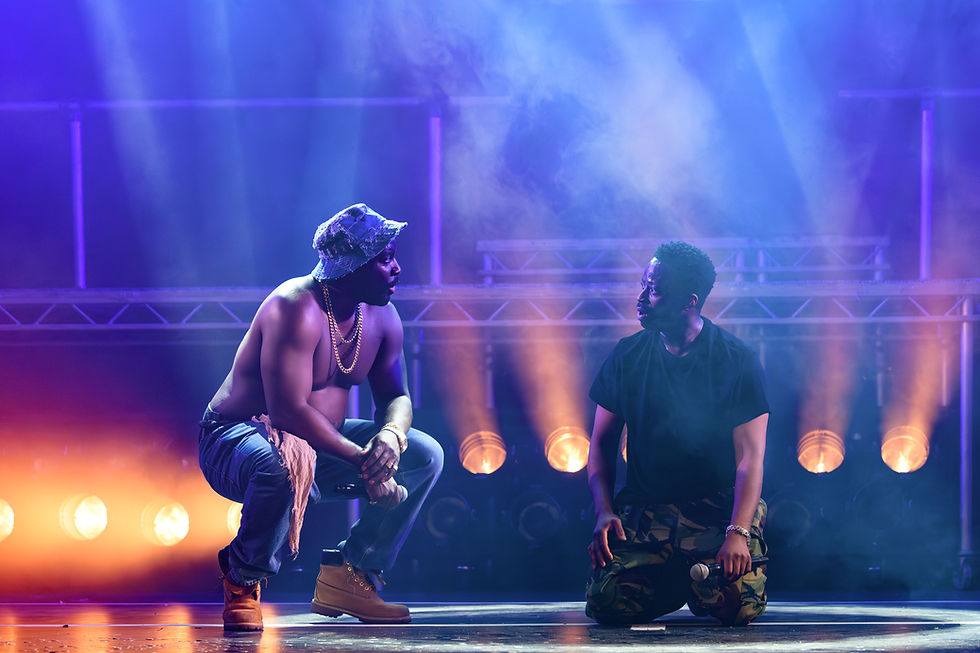 Two performers kneel on stage in dramatic lighting, one shirtless with chains. A moody ambiance with blue and orange lights behind.