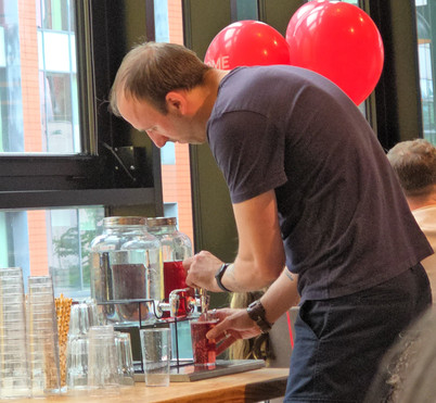 HOME Manchester - Man in dark T-shirt pours a red drink from a glass dispenser at a café table, with red balloons and stacked cups nearby.
