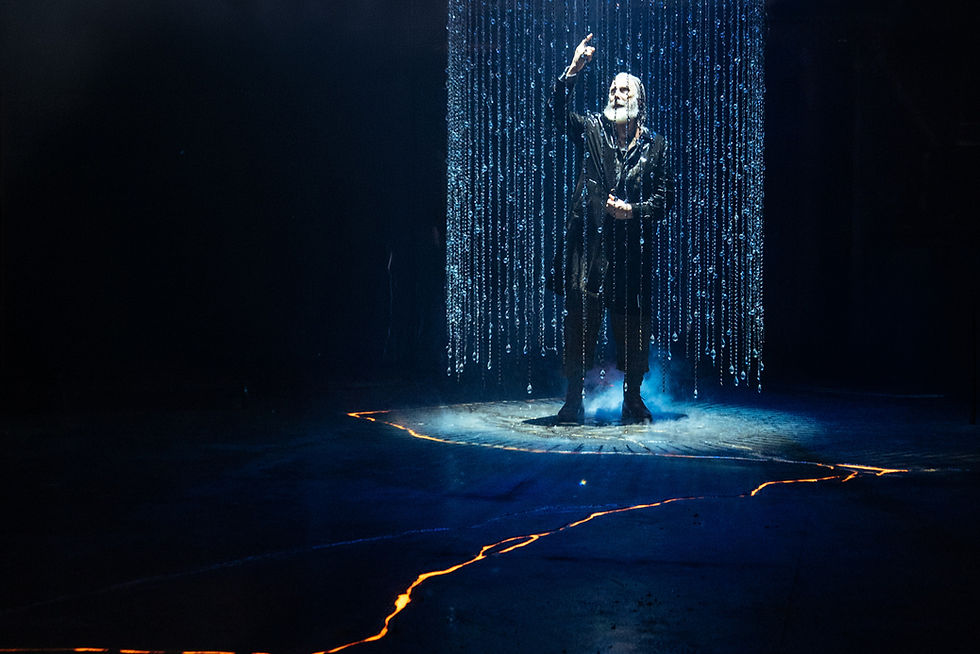 David Threlfall as King Lear standing beneath falling rain, illuminated by blue lighting above a dark stage with glowing gold cracks at the Royal Exchange Theatre.