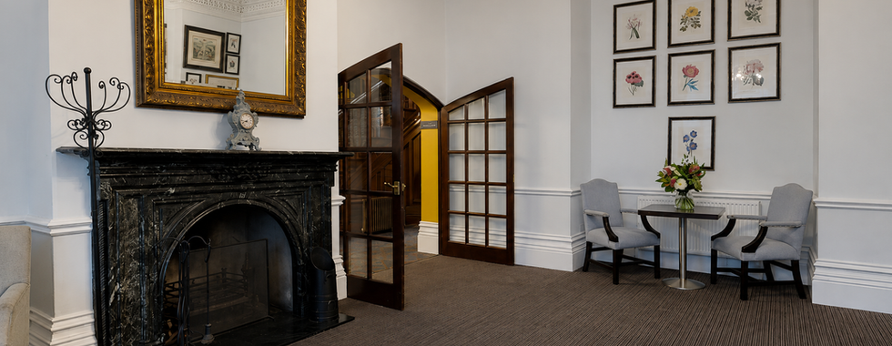 Bright seating area inside Hollin House Hotel featuring a black marble fireplace, decorative gold mirror, floral artwork wall, and two grey armchairs beside a small table with fresh flowers. Wooden glass-panel doors open into a warmly lit corridor.