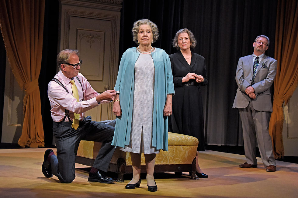 from the play By Royal Appointment - Man kneels, holding tape measure near elderly woman in blue. Two others stand nearby. Elegant room with yellow drapes, formal attire.