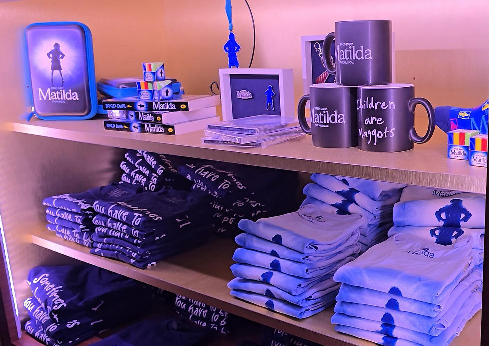 Shelves display Matilda-themed merchandise: books, mugs with text, T-shirts, accessories. Blue and black colors dominate the setting.