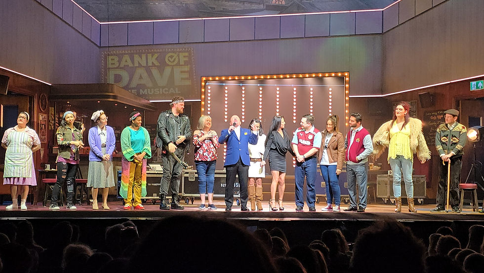Cast bows on stage under Bank of Dave the Musical backdrop, lit by marquee lights, in front of cheering audience.