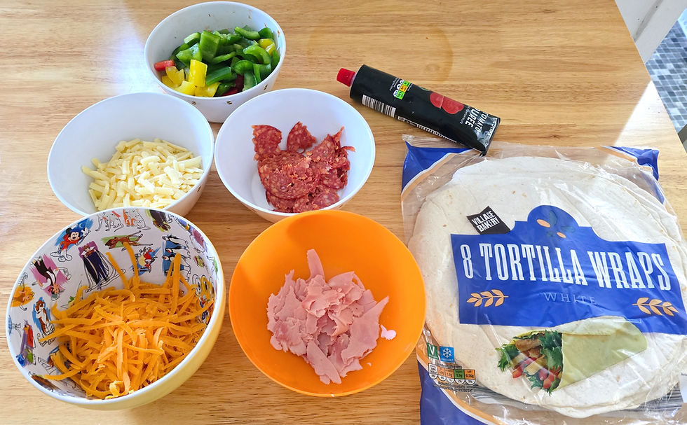 Bowls of sliced peppers, cheese, ham, and salami on a table with tortilla wraps and tomato puree. Bright colors, food prep setting.