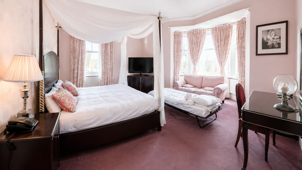 Spacious Brooke Suite bedroom at Hollin House Hotel with a four-poster double bed, pink décor, dressing table, TV stand, pink sofa and camp bed set up by the bay window.