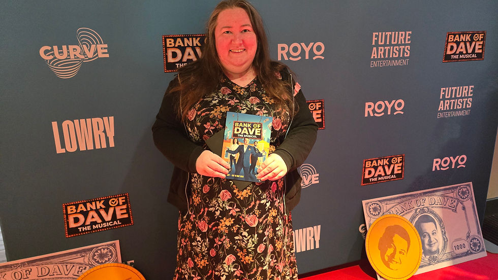 Smiling woman in floral dress holds Bank of Dave the Musical flyer at a branded step-and-repeat backdrop.