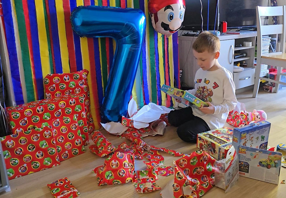 Child opening gifts at a birthday party with a blue "7" balloon and Mario-themed decorations. Wrapped presents scattered on the floor.