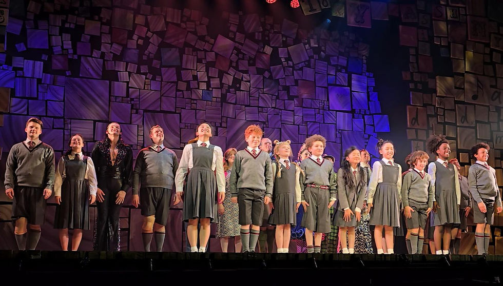 Stage performance with a group of people in school uniforms, smiling under colorful spotlights, against a backdrop of patterned tiles.