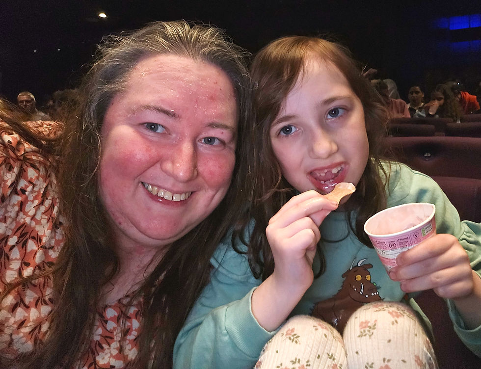 Smiling woman and child eating ice cream in a theater, seated with others. The child wears a green shirt with a Gruffalo design.
